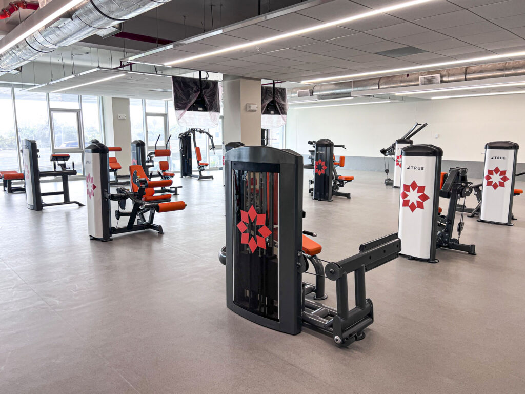 A spacious, modern gym with various exercise machines featuring orange seats and white panels with red star logos, large windows letting in natural light, and a clean, organized layout.