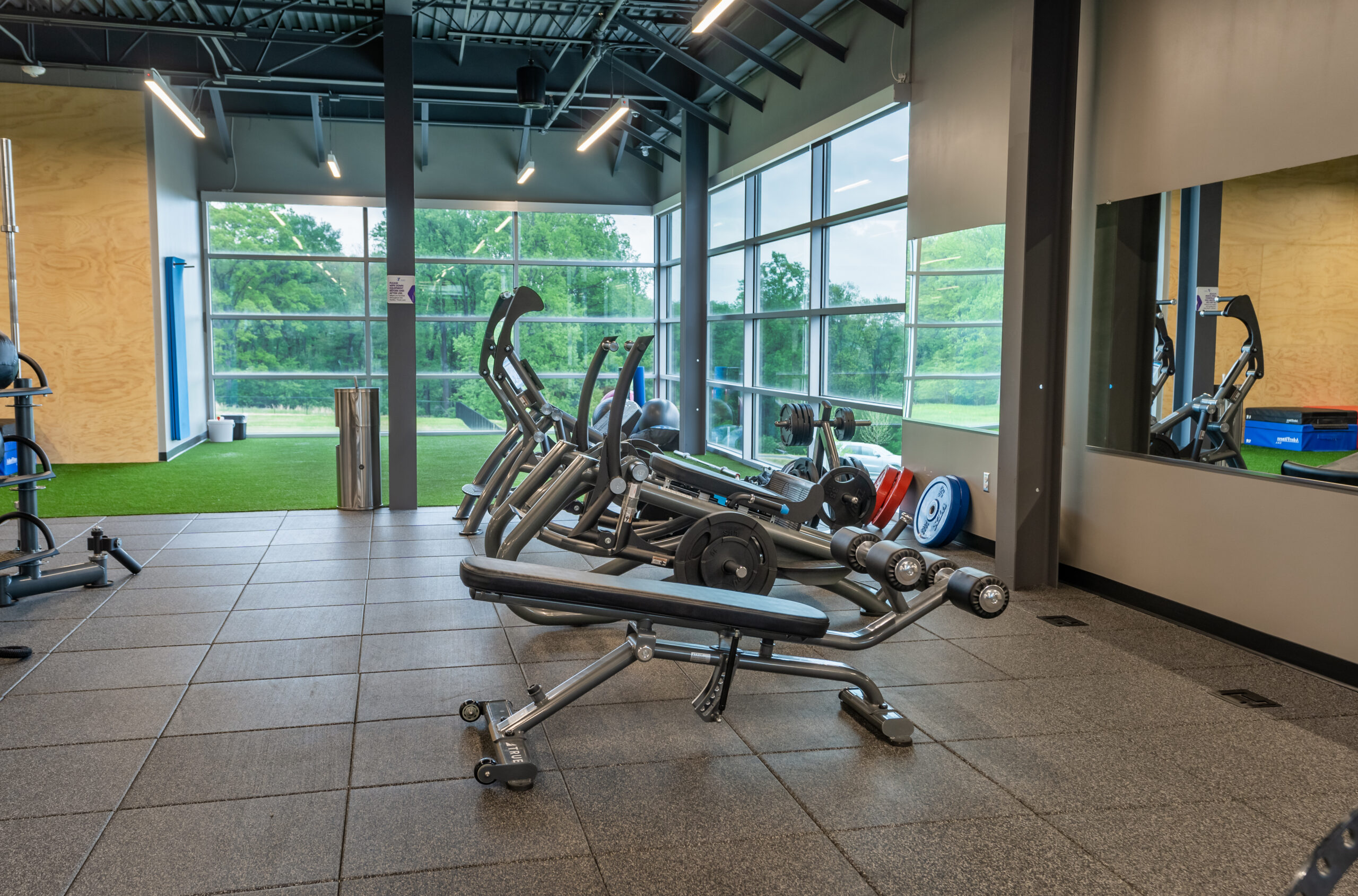 A modern gym with weight benches, dumbbells, and cardio machines near large windows, overlooking greenery. The space has gray floors, high ceilings, and a mirrored wall on the right.