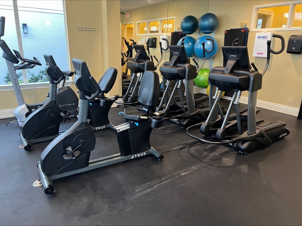 A gym room with two stationary exercise bikes and three elliptical machines, all facing mirrors. There are three colorful exercise balls stacked in the corner. The room has a window letting in natural light.
