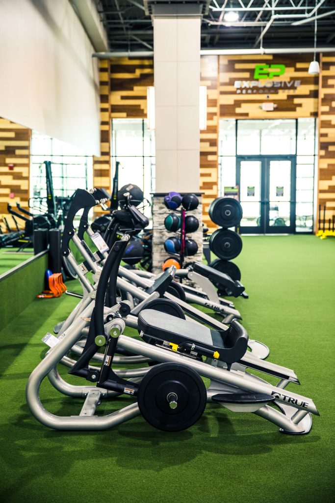 Several elliptical machines are lined up on green turf inside a modern gym. Medicine balls and weight plates are stored nearby, with natural light streaming through large windows in the background.
