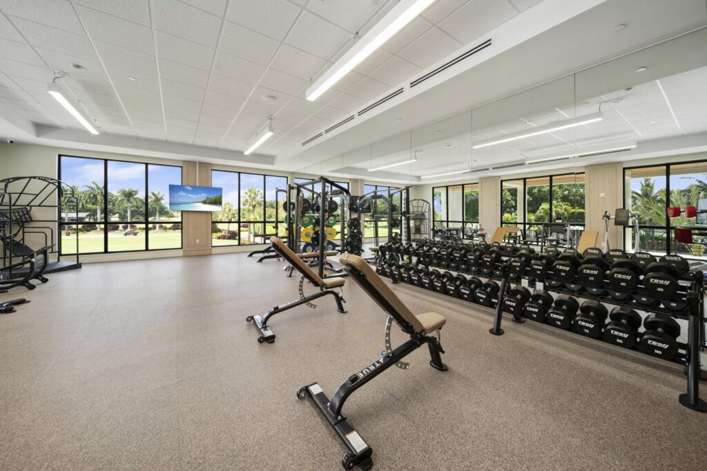 A spacious gym with large windows, weight racks, benches, mirrors, cardio machines, and a view of palm trees and greenery outside. The room is bright with ample natural light.