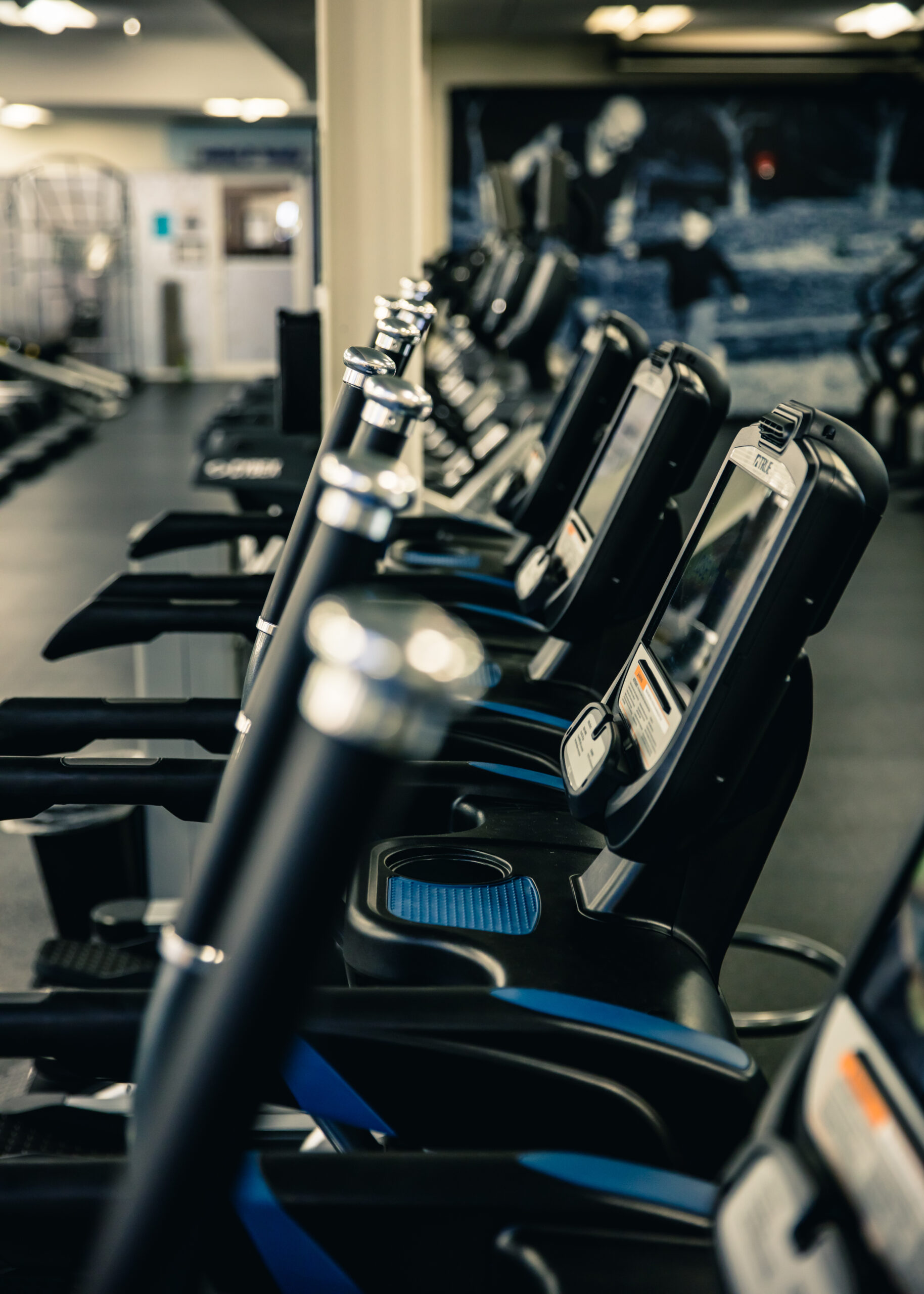 A row of stationary exercise bikes with digital screens lined up inside a gym, with other gym equipment visible in the blurred background.