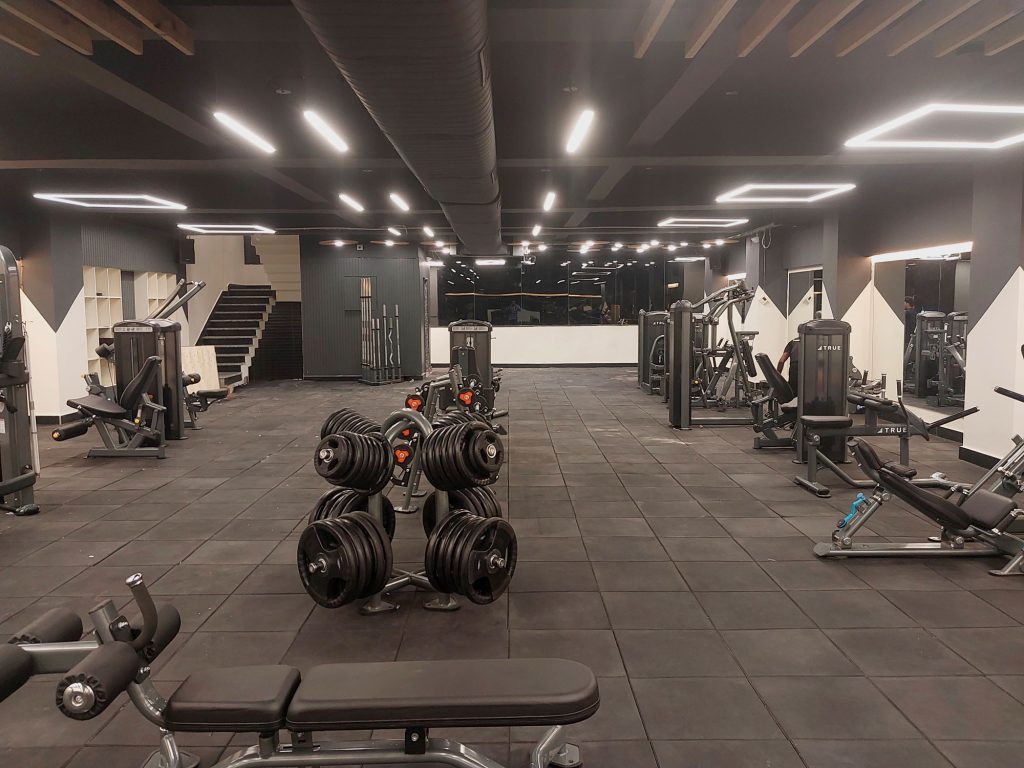Modern gym with black rubber flooring, various exercise machines, weight racks, and benches. The space is well-lit with LED ceiling lights and has a clean, organized appearance. No people are present.