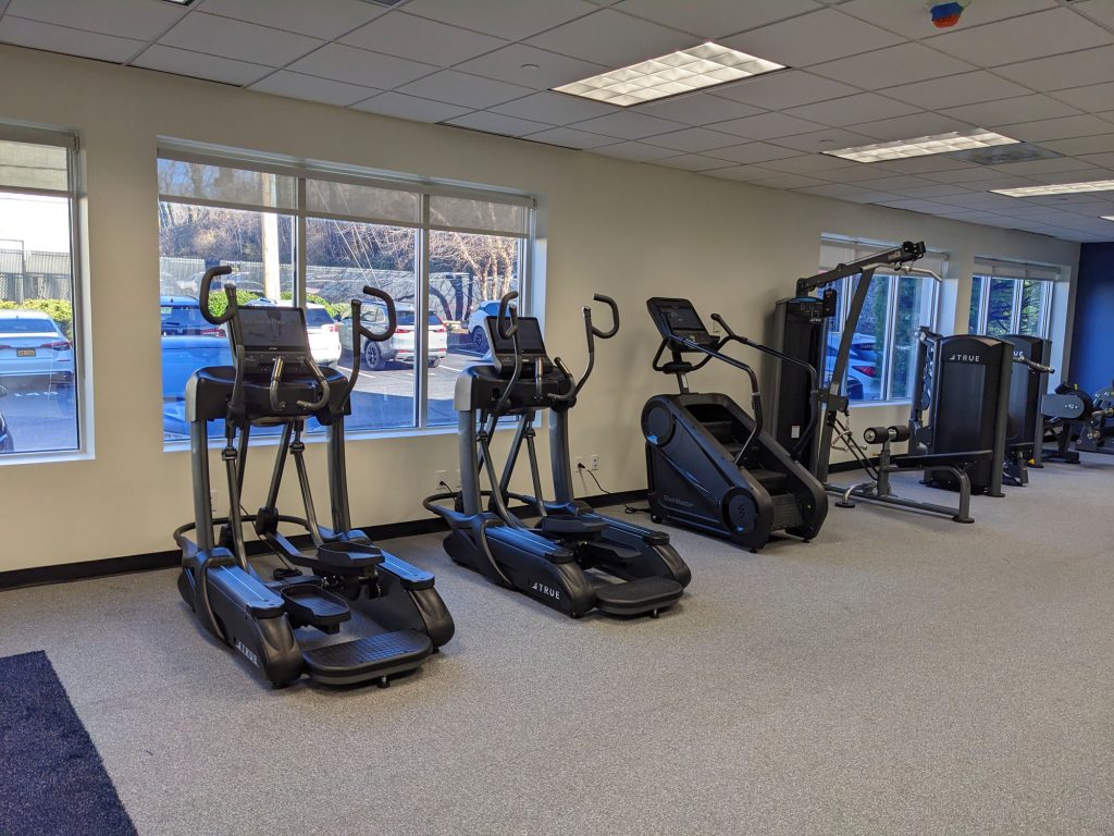 A gym with large windows showing parked cars outside, featuring two elliptical machines, a stair climber, and strength training equipment on a light-colored carpet.