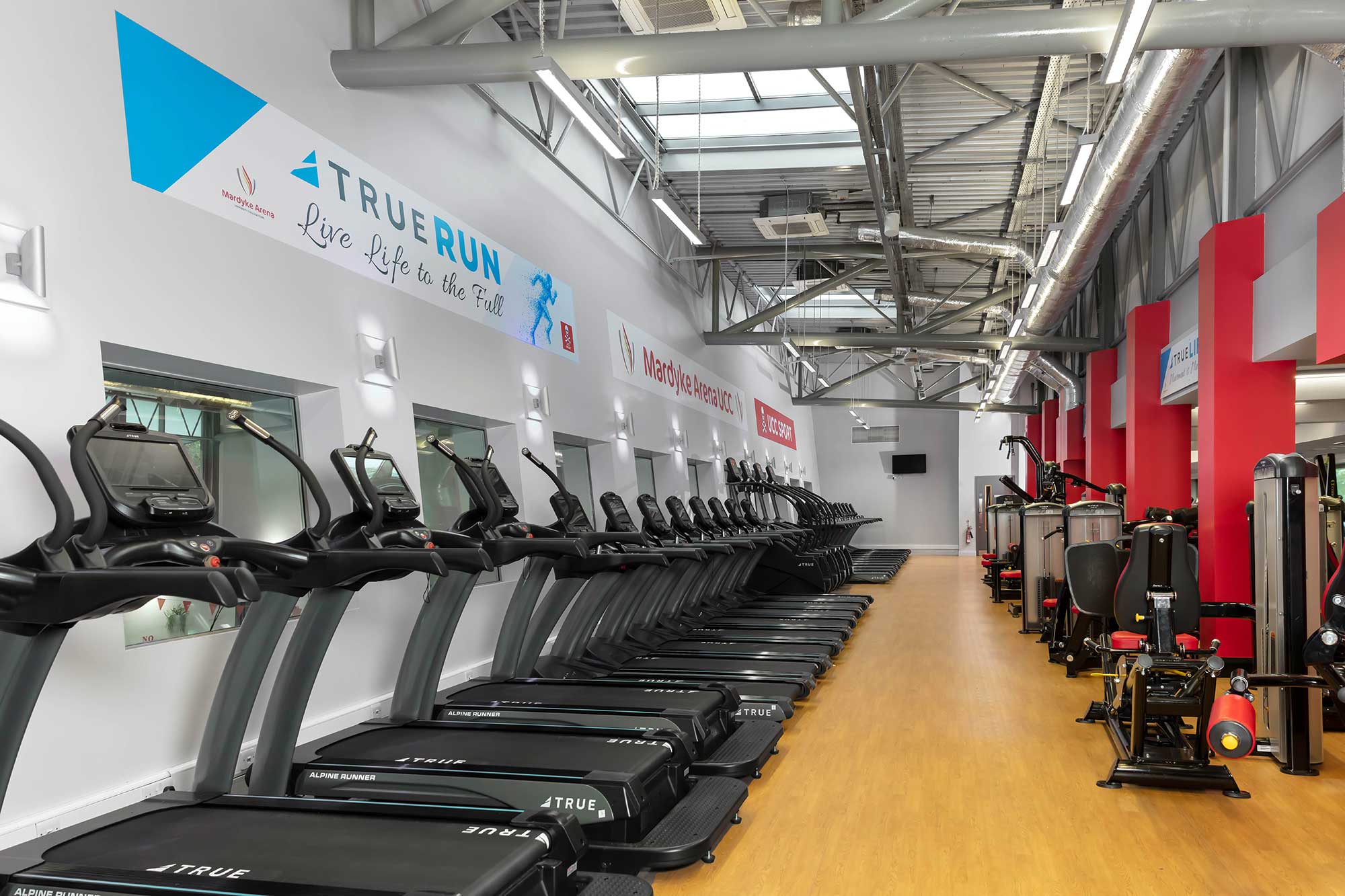 A modern gym with a row of treadmills lined up along the left wall, strength training machines on the right, bright lighting, wood flooring, and motivational banners on the white and red walls.