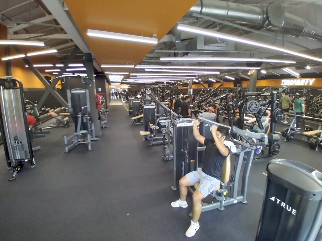 A spacious gym with people using various exercise machines. One person in the foreground is seated on a chest press machine, lifting weights. The gym is well-lit with orange and black accents and has modern fitness equipment.
