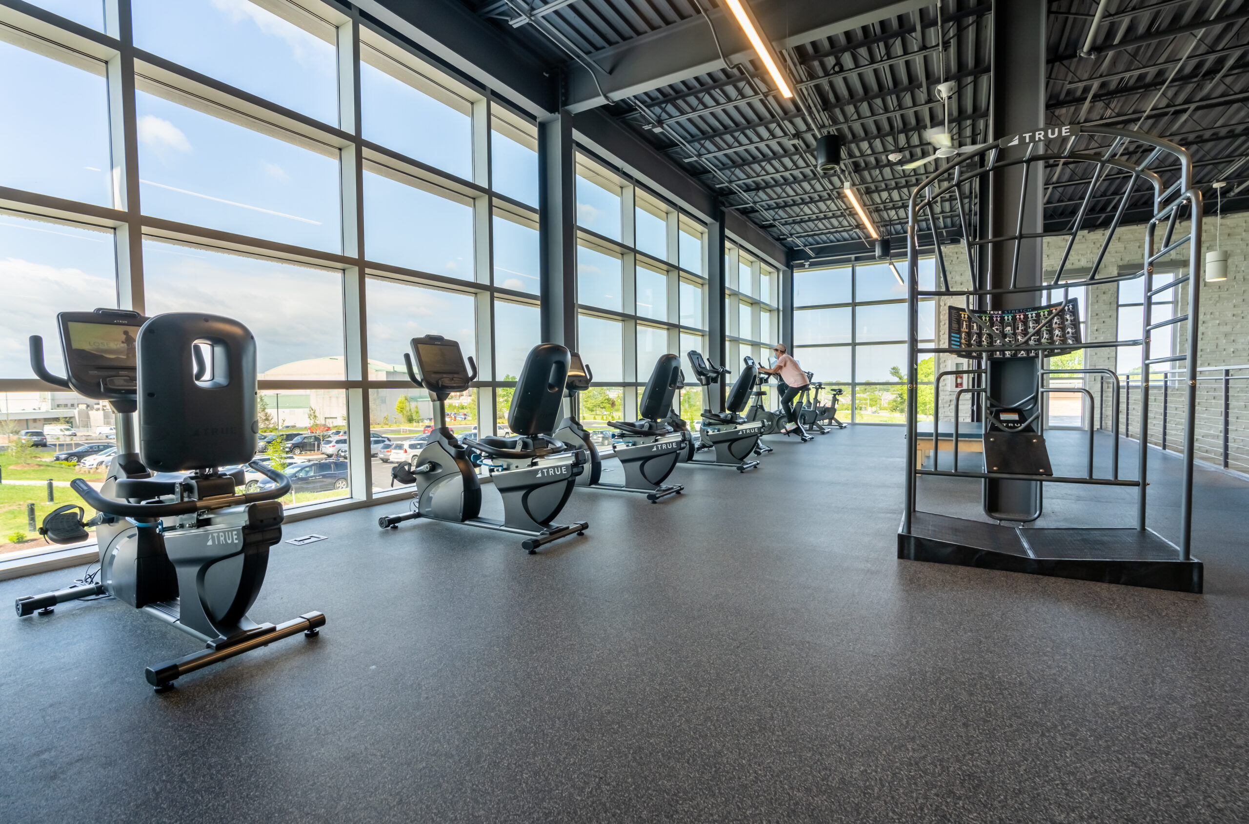 A modern gym with large windows, several stationary exercise bikes lined up, and a cardio climbing machine, all in a bright, spacious room with outdoor views.