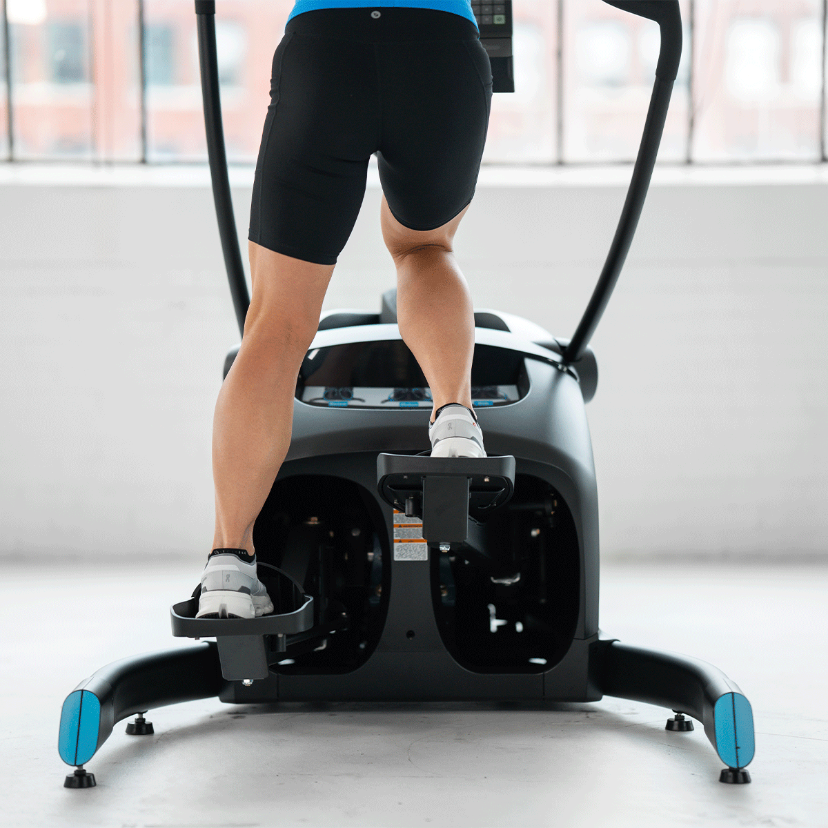 A person exercising on an elliptical machine in a bright room, seen from behind, wearing black shorts and sneakers. Large windows are visible in the background.