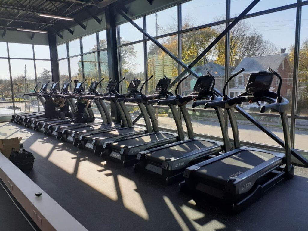 A row of treadmills lines a large window in a bright, modern gym with sunlight streaming in. Some equipment and boxes are on the floor, and houses and trees are visible outside.
