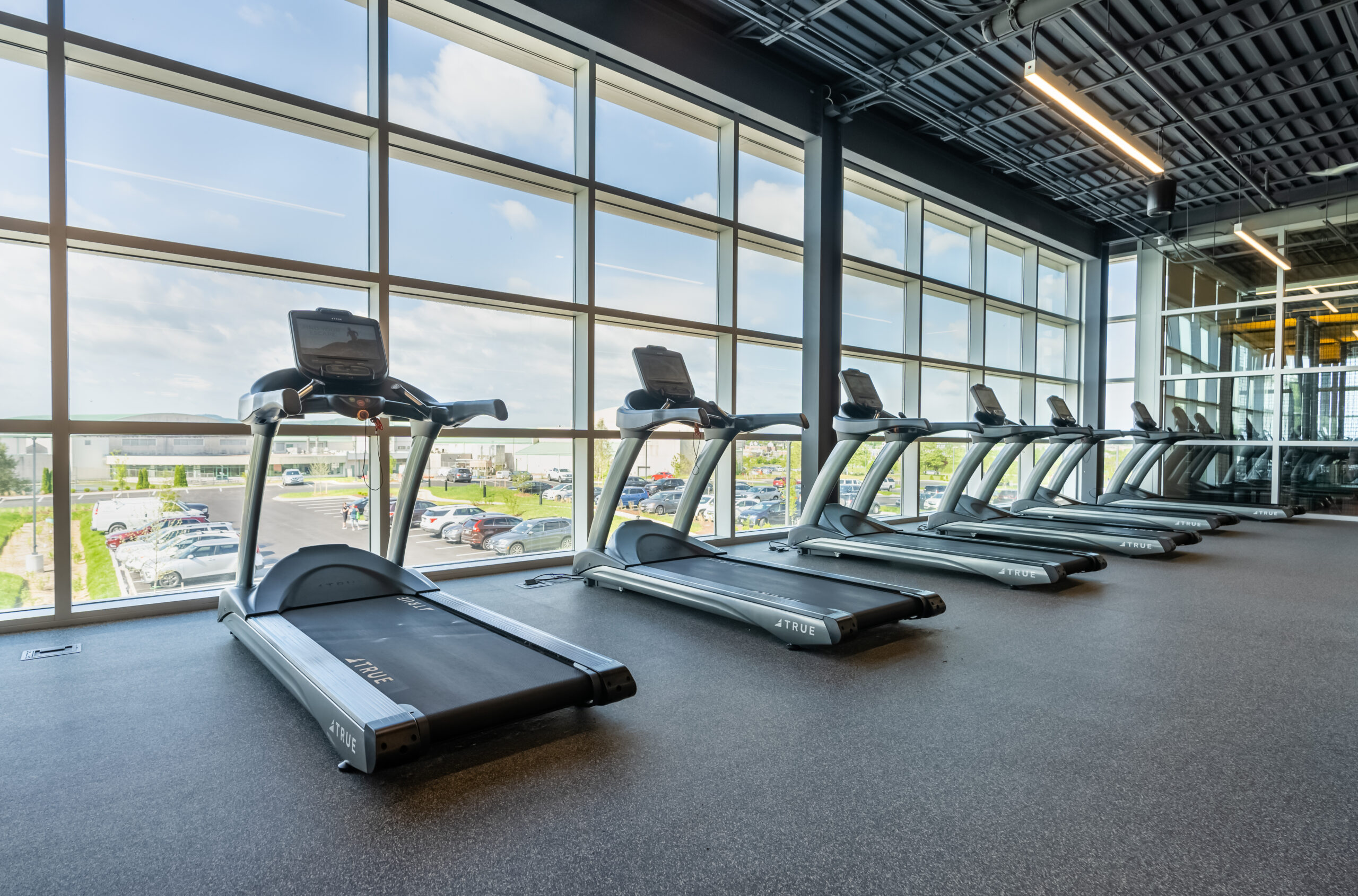 A row of treadmills lined up in a modern gym with large floor-to-ceiling windows, allowing natural light to fill the space. Cars and buildings are visible outside through the windows.