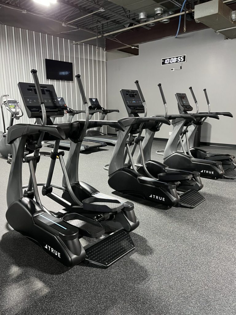 Three elliptical machines lined up in a modern gym with gray walls, a digital clock on the wall, and other fitness equipment visible in the background. The floor is covered with dark speckled rubber.