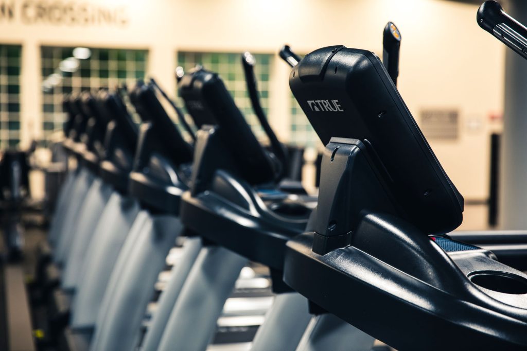 A row of black and gray treadmills in a gym, lined up side by side. The focus is on one treadmill console in the foreground, with the others gradually fading into the background.