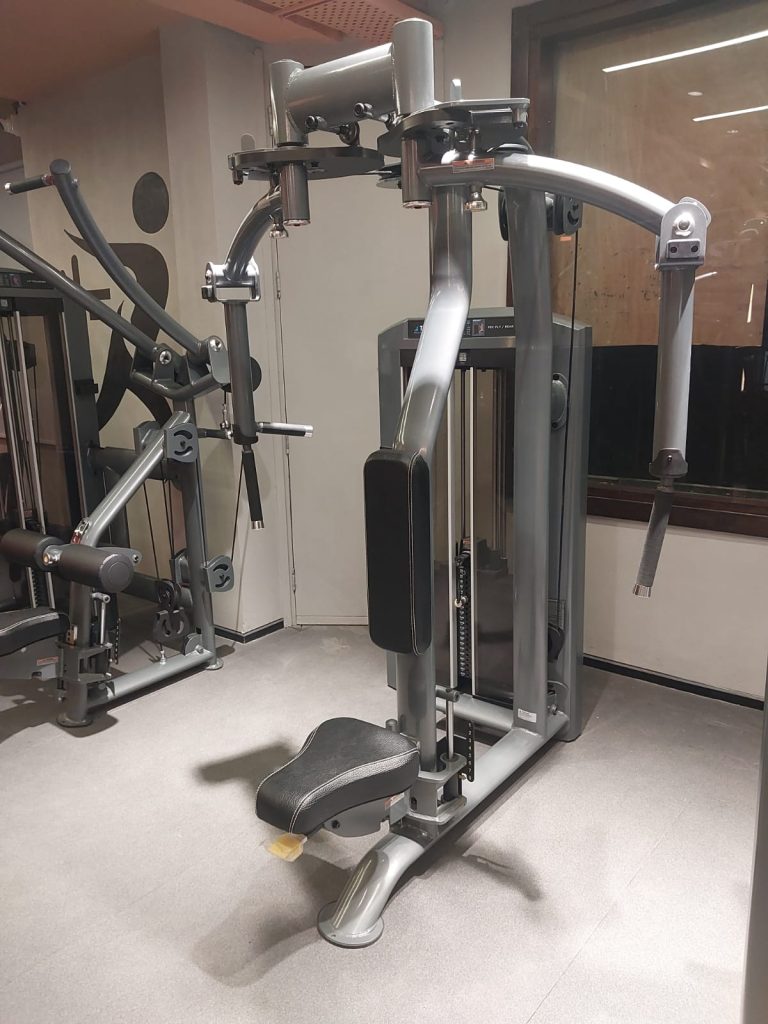 A pec deck machine in a gym, featuring a padded seat and backrest with two adjustable arms and handles for chest fly exercises. The machine is metallic gray and set against a neutral wall and window.