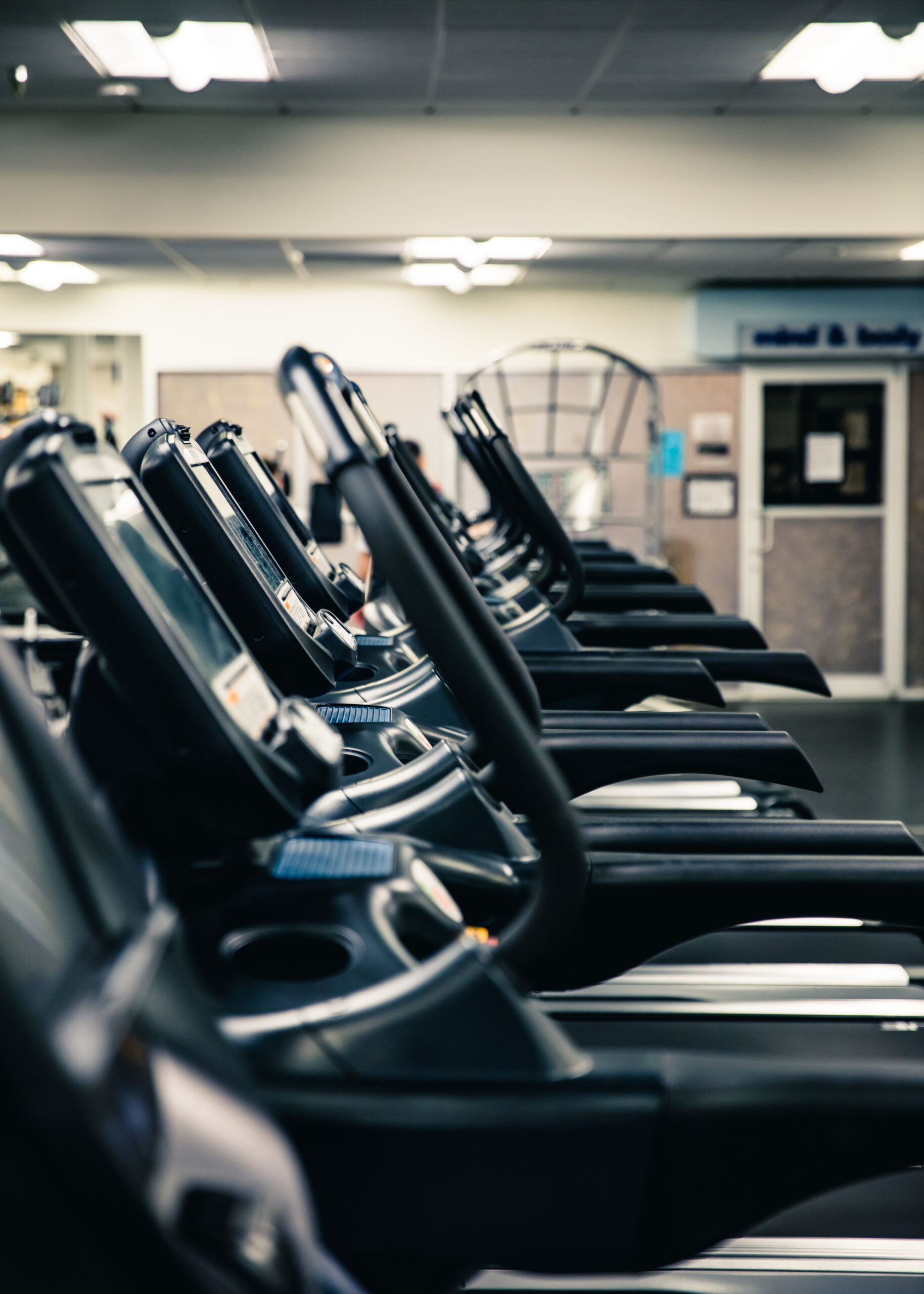 A row of treadmills lined up inside a gym, with a blurred background showing gym equipment and fluorescent ceiling lights.