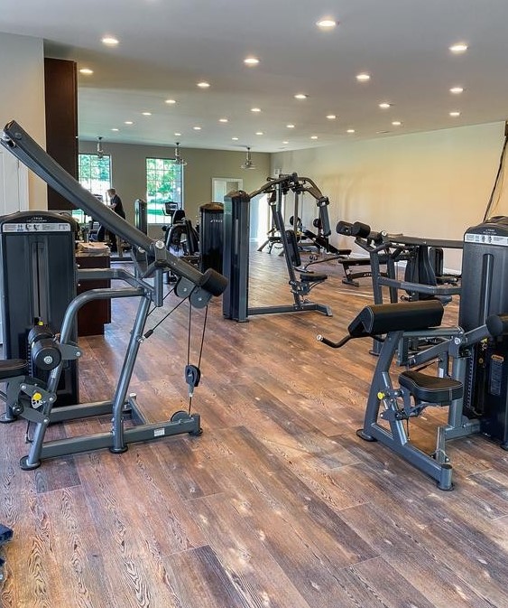 A spacious gym with wooden floors, large windows, and various exercise machines including cable machines, elliptical trainers, and leg curl machines, all neatly arranged in a well-lit room.