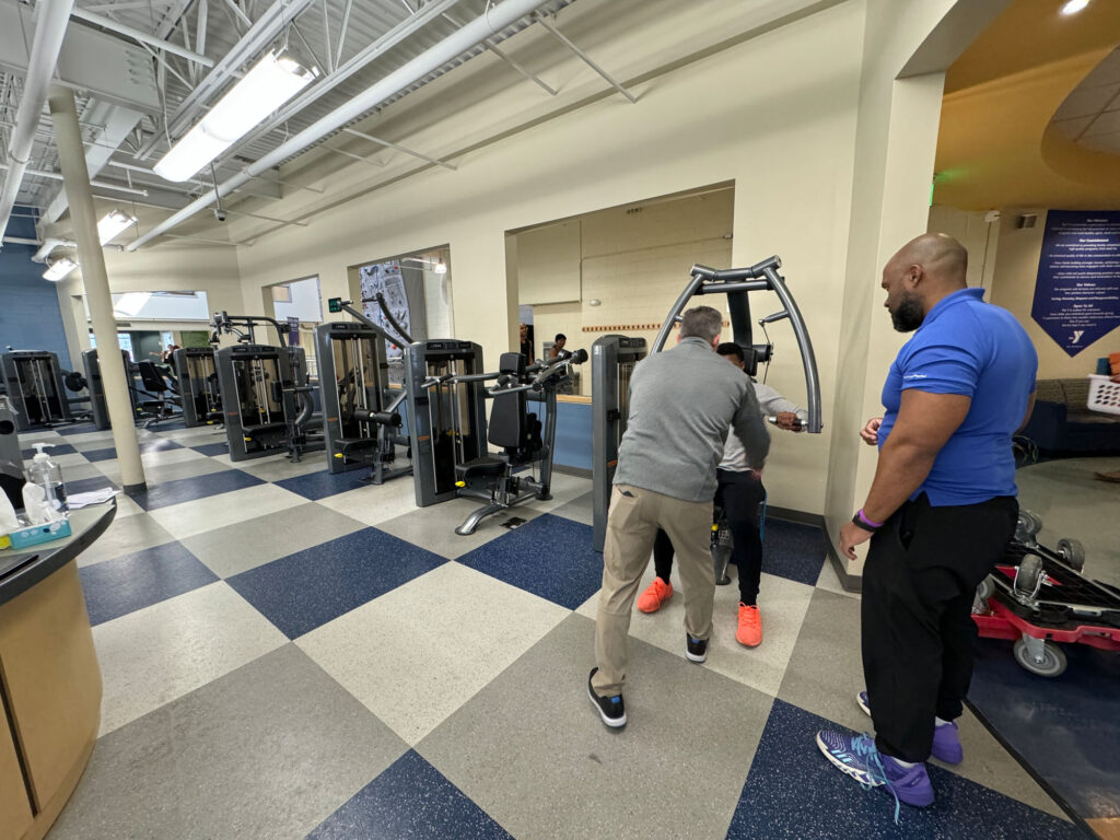 Two men assist a third man using a strength training machine in a spacious, well-lit gym. Other exercise equipment is visible in the background, and the floors have a blue and gray checkered pattern.