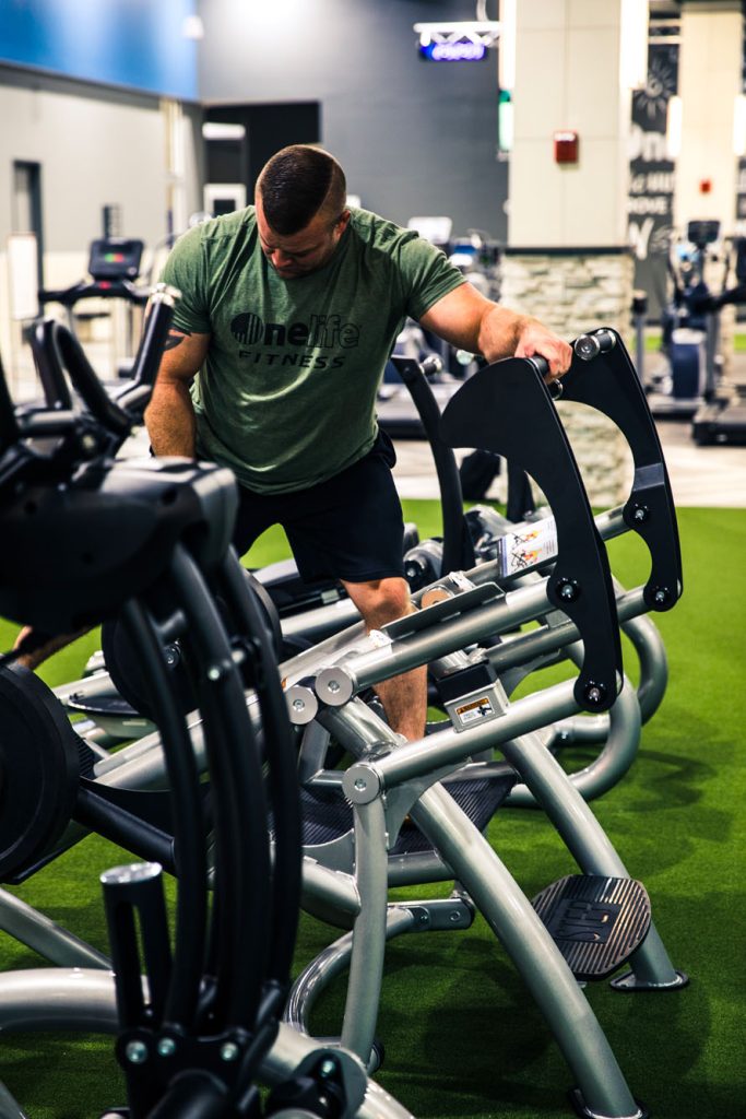 A man wearing a green Onelife Fitness shirt adjusts weights on a gym machine in a spacious, modern fitness center with green artificial turf flooring.