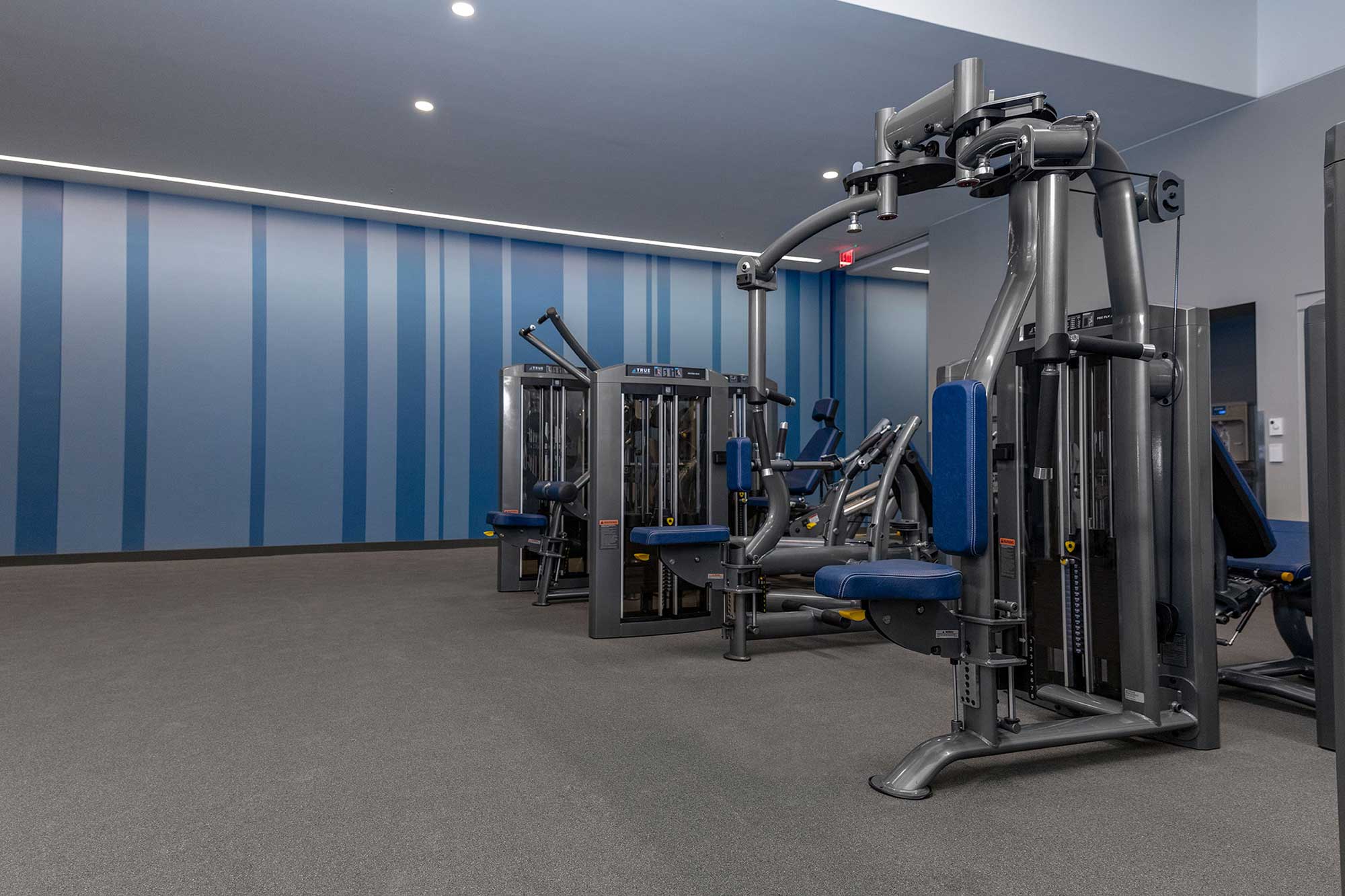 A modern gym room with several weight machines featuring blue seats and padding. The walls are decorated with vertical blue stripes, and the floor is covered with gray carpet. The room is well-lit and spacious.
