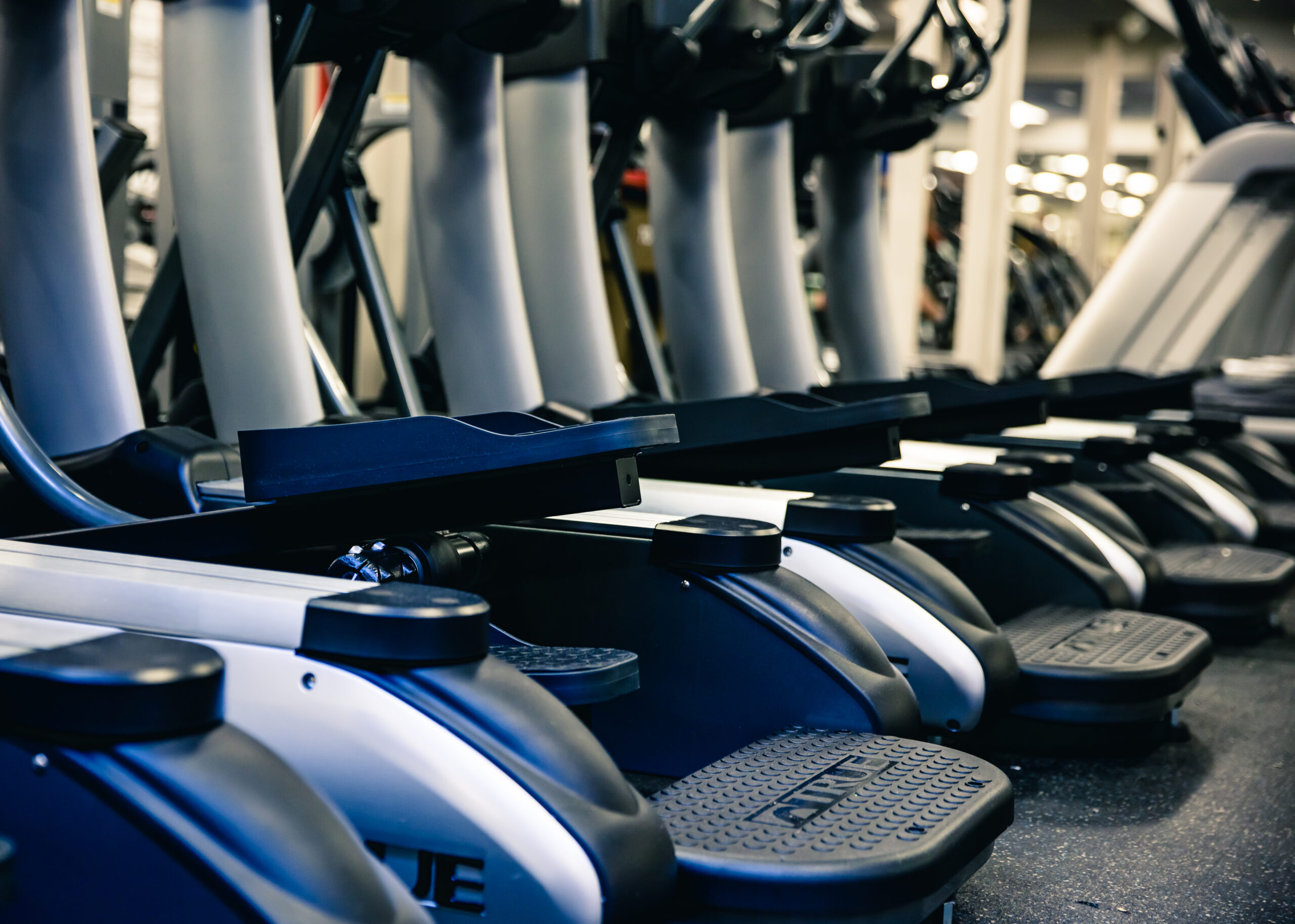 Row of elliptical machines at a gym, closely lined up with foot pedals and handles visible, in a brightly lit indoor setting.