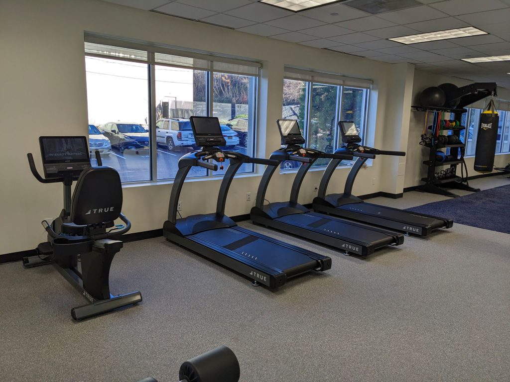 A gym with a stationary bike, three treadmills, and a rack holding exercise balls, kettlebells, and weights near large windows overlooking a parking lot.