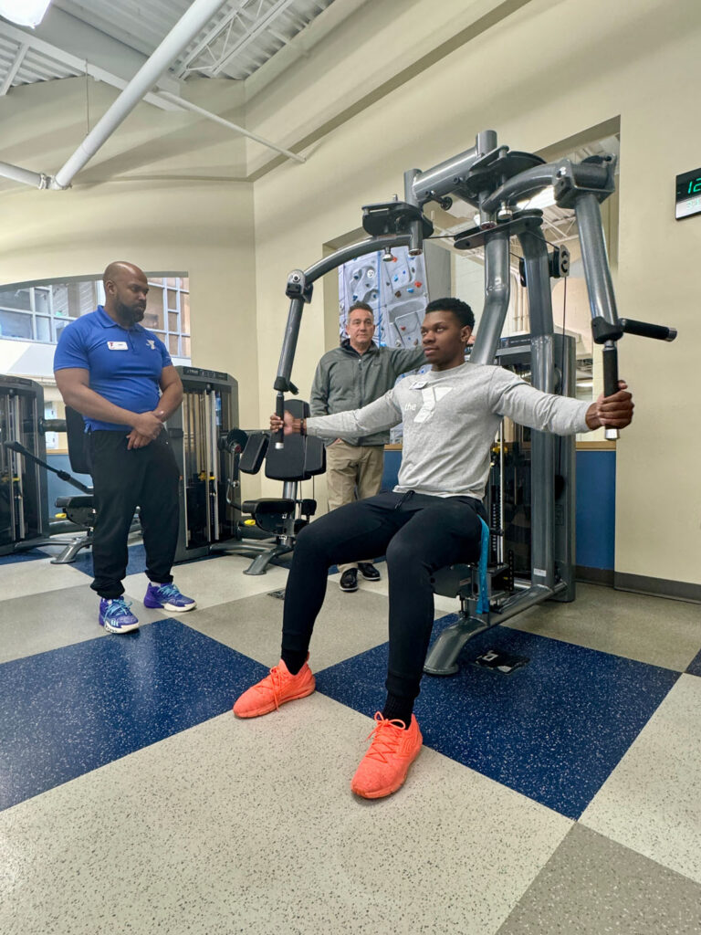 A young man uses a chest fly machine at the gym while two men observe; one stands nearby in a blue shirt, and the other stands in the background. The gym has blue and white tiled floors.