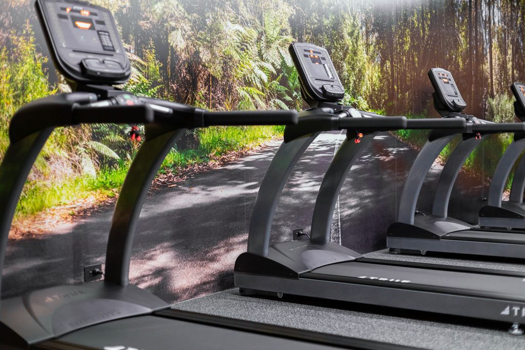 Several treadmills are lined up indoors, facing a wall mural that depicts a sunlit forest path, creating an outdoor running illusion in the gym.