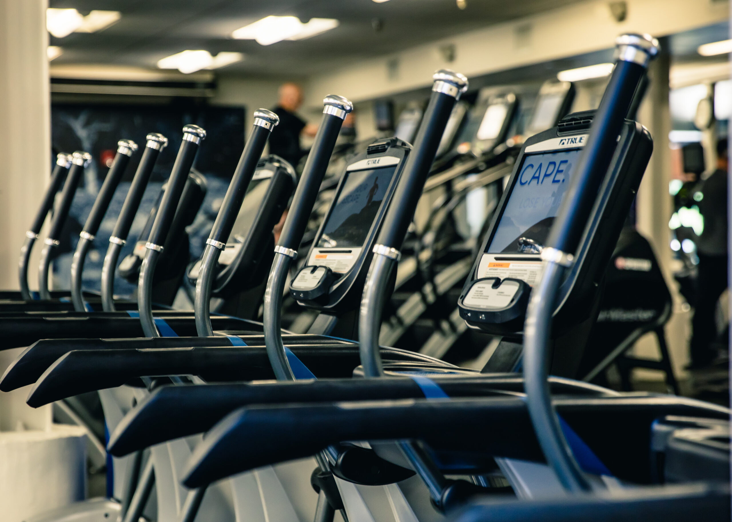 A row of elliptical machines lined up in a gym, with screens and handles visible, under bright overhead lighting and blurred background showing mirrors and gym equipment.