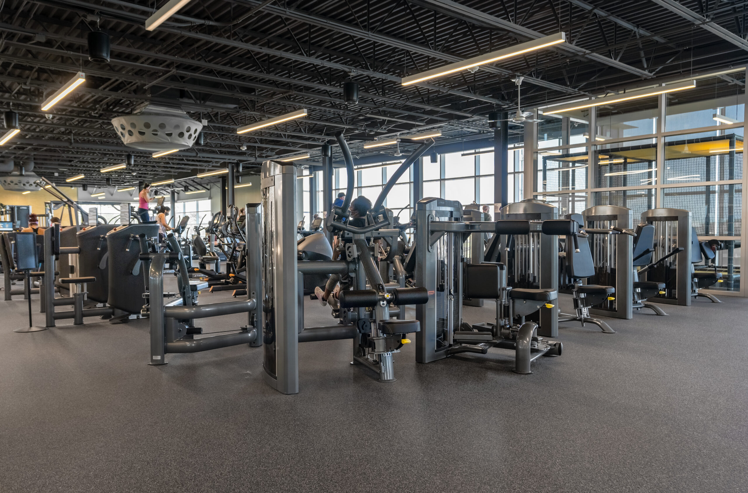 A spacious gym with various exercise machines, including weight equipment and cardio machines, set up in a bright room with large windows and exposed ceiling. Few people are working out in the background.