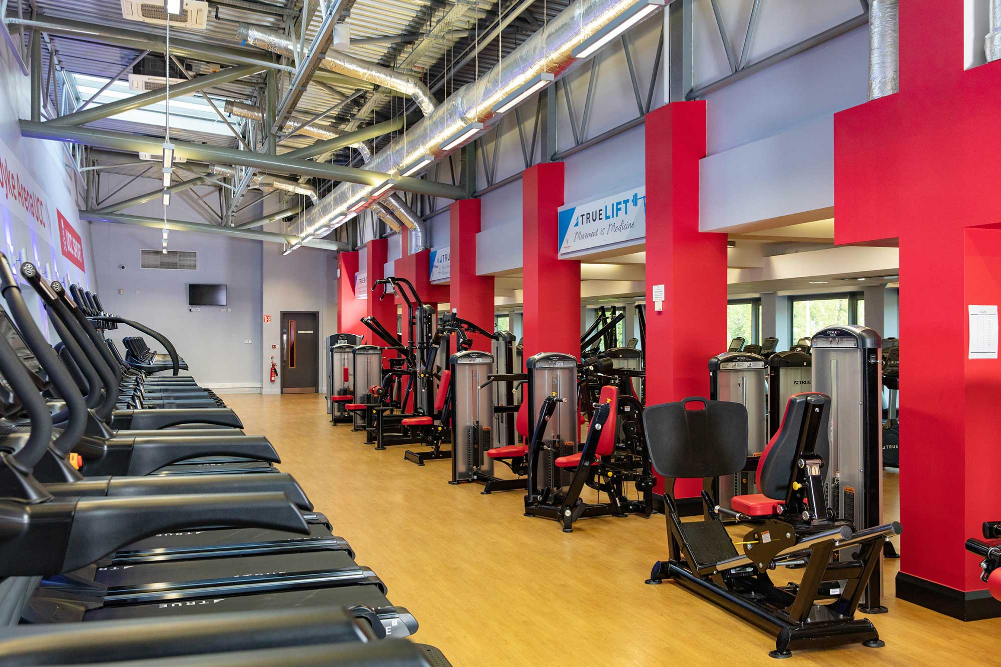 A modern gym interior with rows of treadmills on the left and various strength training machines with red seats on the right, set on a wooden floor with exposed ductwork on the ceiling.