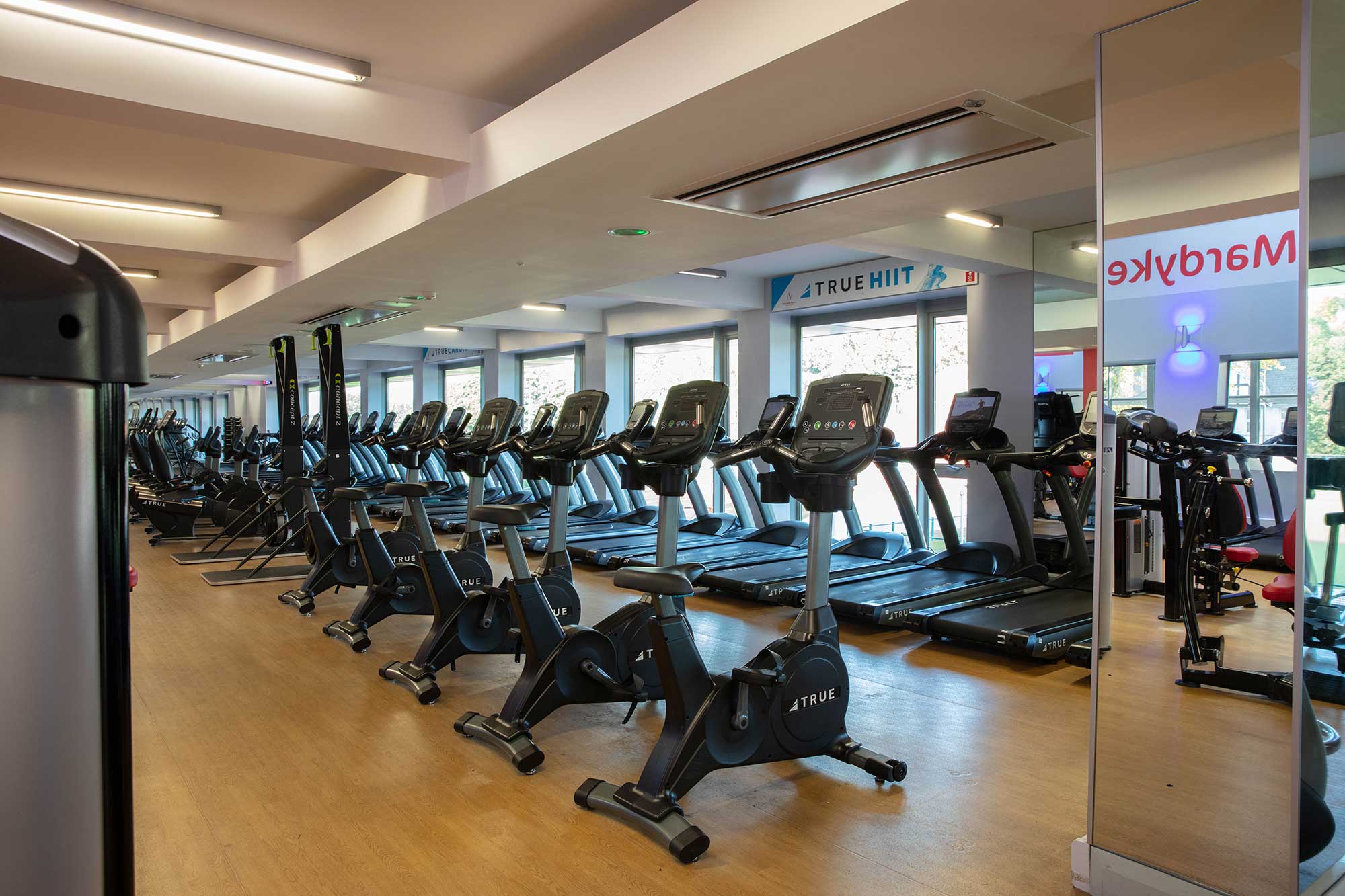 A gym with rows of treadmills and exercise bikes on a wooden floor, large mirrors on one wall, and windows letting in natural light.