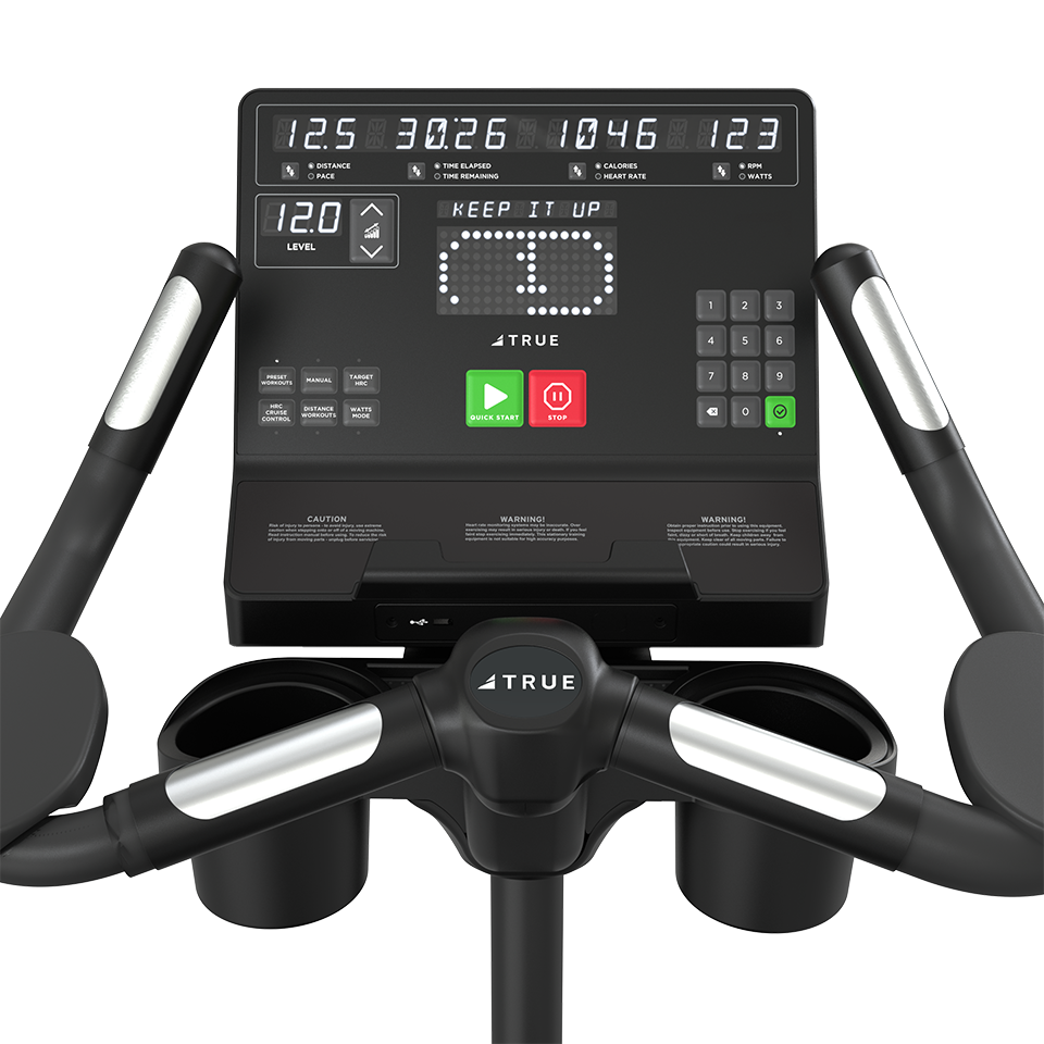Close-up of a treadmill console displaying workout information such as time, distance, calories, and heart rate. The interface includes buttons, cup holders, and handlebars with the brand name TRUE visible.