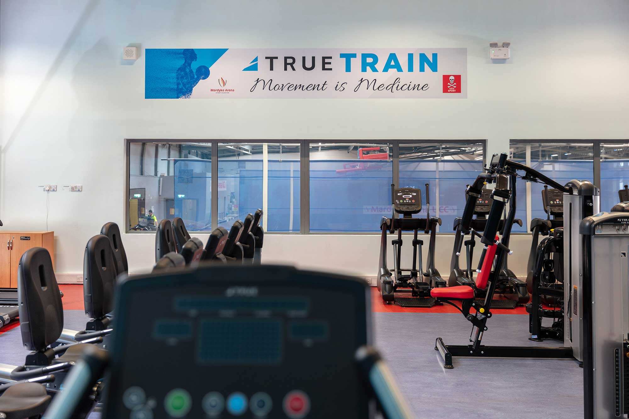 A gym with various exercise machines and a sign on the wall that reads TRUE TRAIN Movement is Medicine above large windows. The focus is on workout equipment, including treadmills and weight machines.