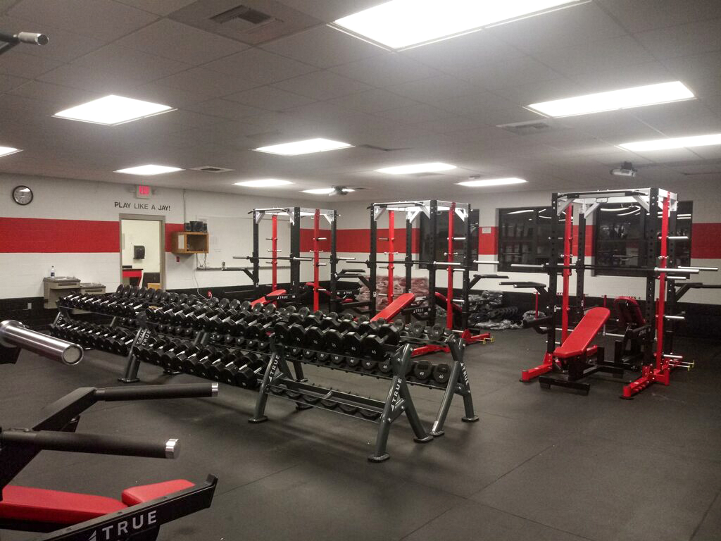 A well-lit gym with rows of dumbbells, weight benches, and red-and-black power racks on a black rubber floor. Mirrors line the wall, and motivational text reads PLAY LIKE A JAY! in the back.
