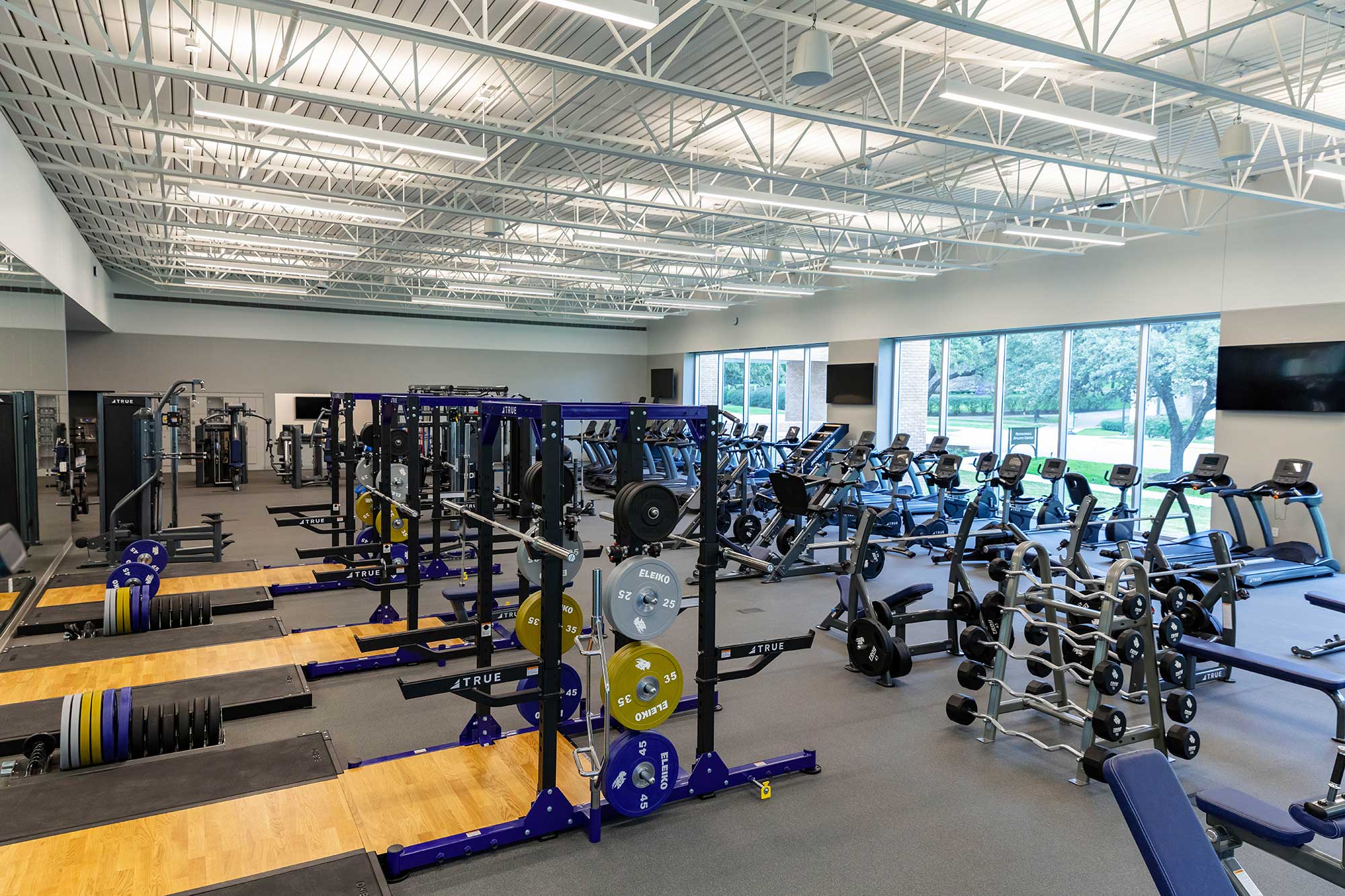 A spacious, modern gym with weightlifting platforms, racks of weights, benches, cardio machines, and large windows letting in natural light. The gym is clean, well-lit, and has various fitness equipment neatly arranged.