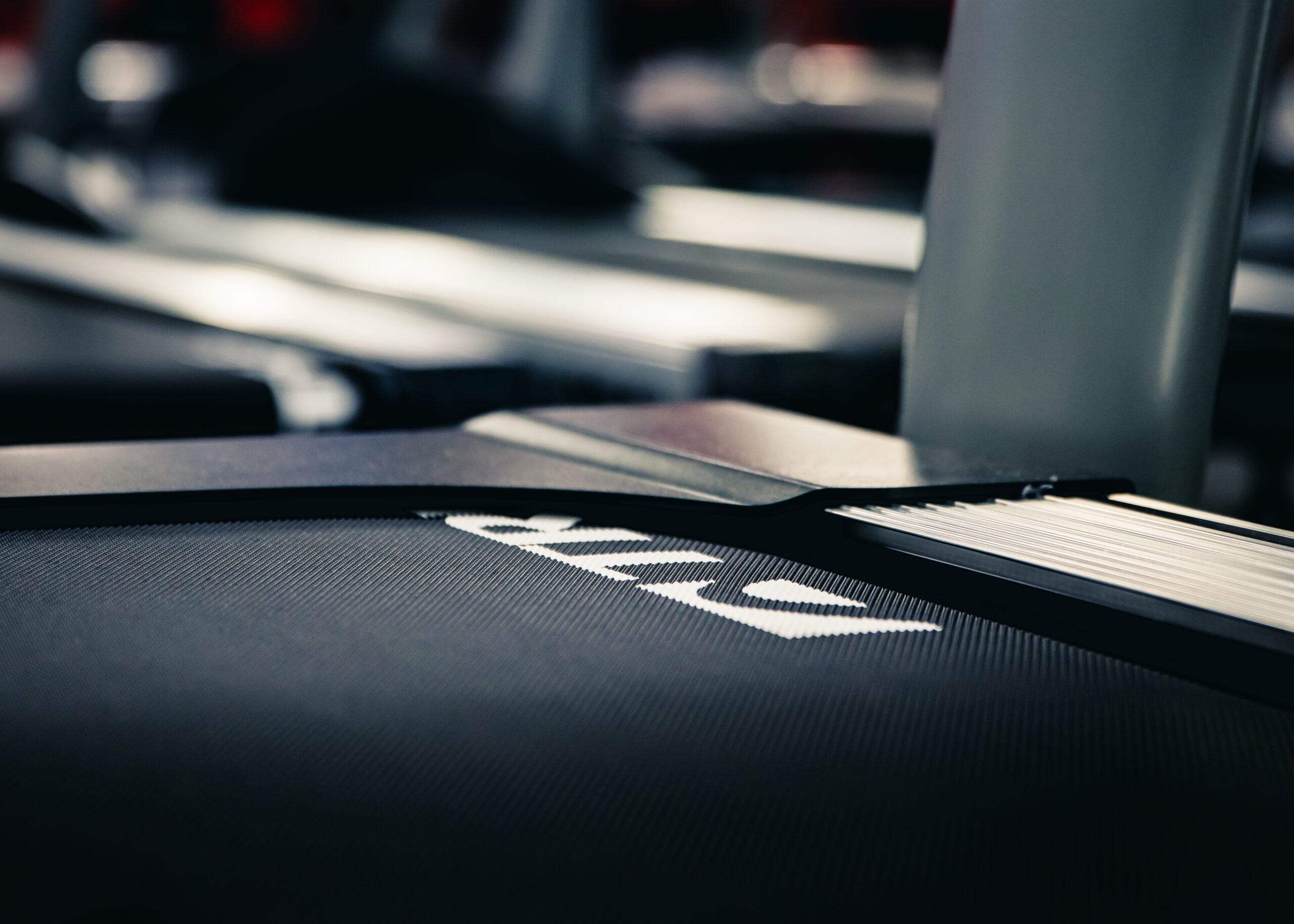 Close-up of a treadmills running belt and display area, with the focus on the textured surface and partially visible white logo or lettering. Background is blurred, emphasizing the equipment detail.