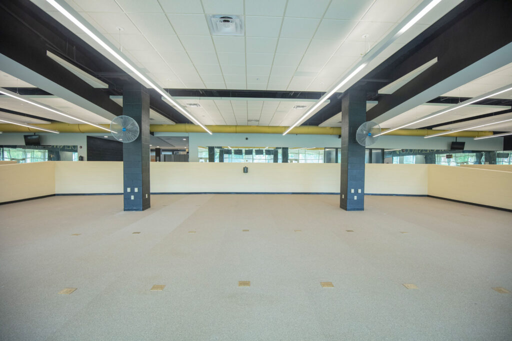 A spacious, empty room with light-colored carpet, high ceilings, fluorescent lights, and black beams. Large windows line the back wall, and there are two black columns in the center of the space.