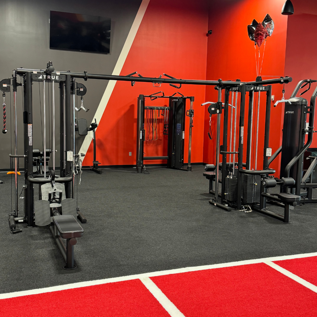 A modern gym with red and gray walls, featuring various strength training machines, cable machines, and exercise equipment on black carpeted flooring. Balloons are visible in the back corner.