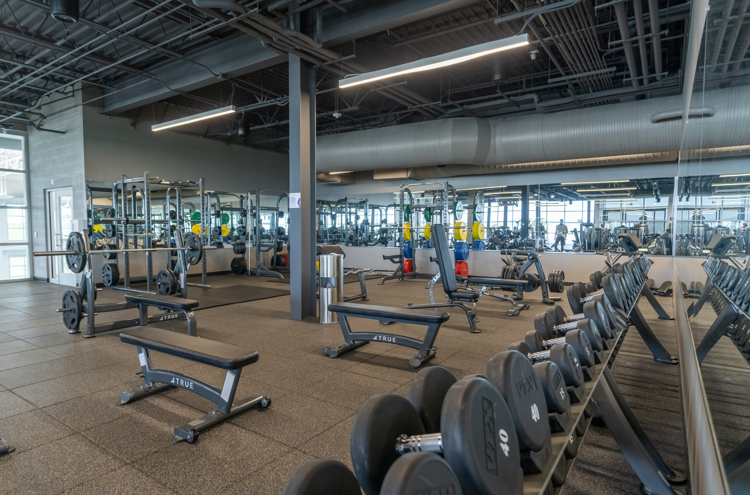 A spacious gym with weight benches, racks of dumbbells, squat racks, and mirrored walls. The area has rubber flooring, industrial-style ceilings, and ample natural light from large windows.