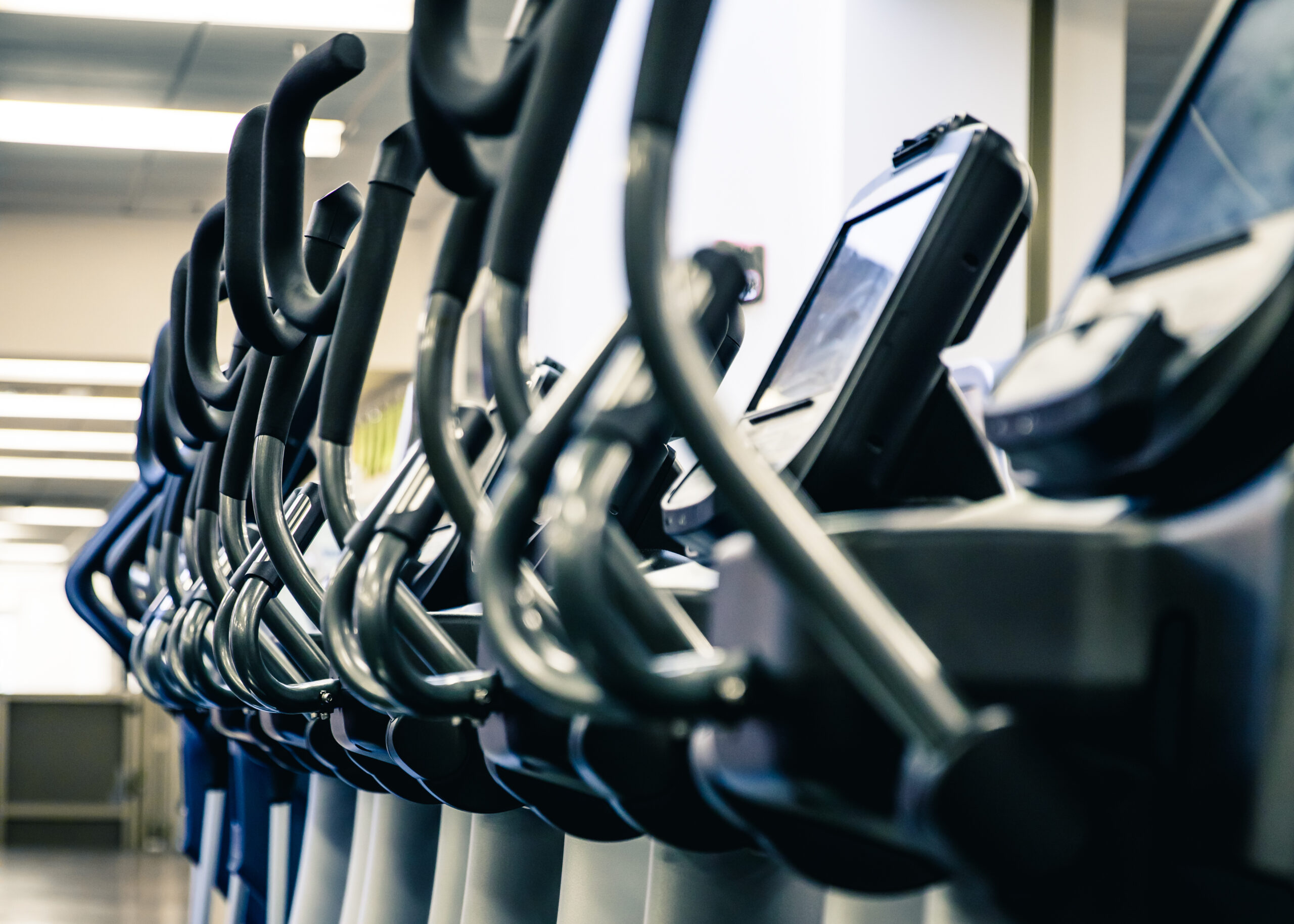A row of exercise bikes or elliptical machines is lined up in a gym, viewed from the side, with digital screens and handlebars in focus. The background shows bright, indoor lighting.