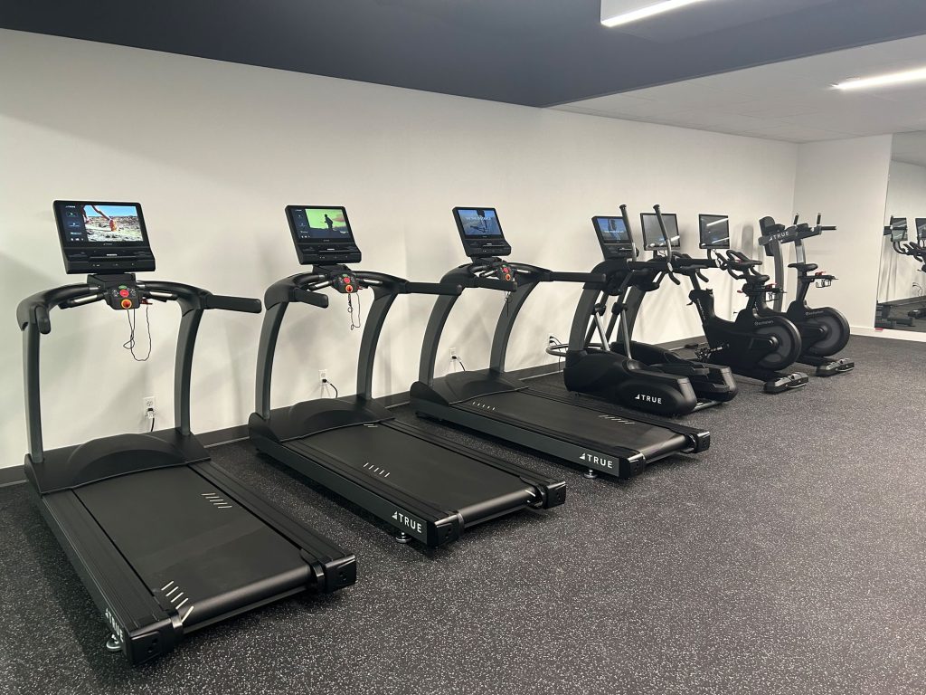 A gym with three treadmills and three elliptical machines lined up against a white wall, each with a digital display, on a black speckled floor. The space is well-lit and modern.
