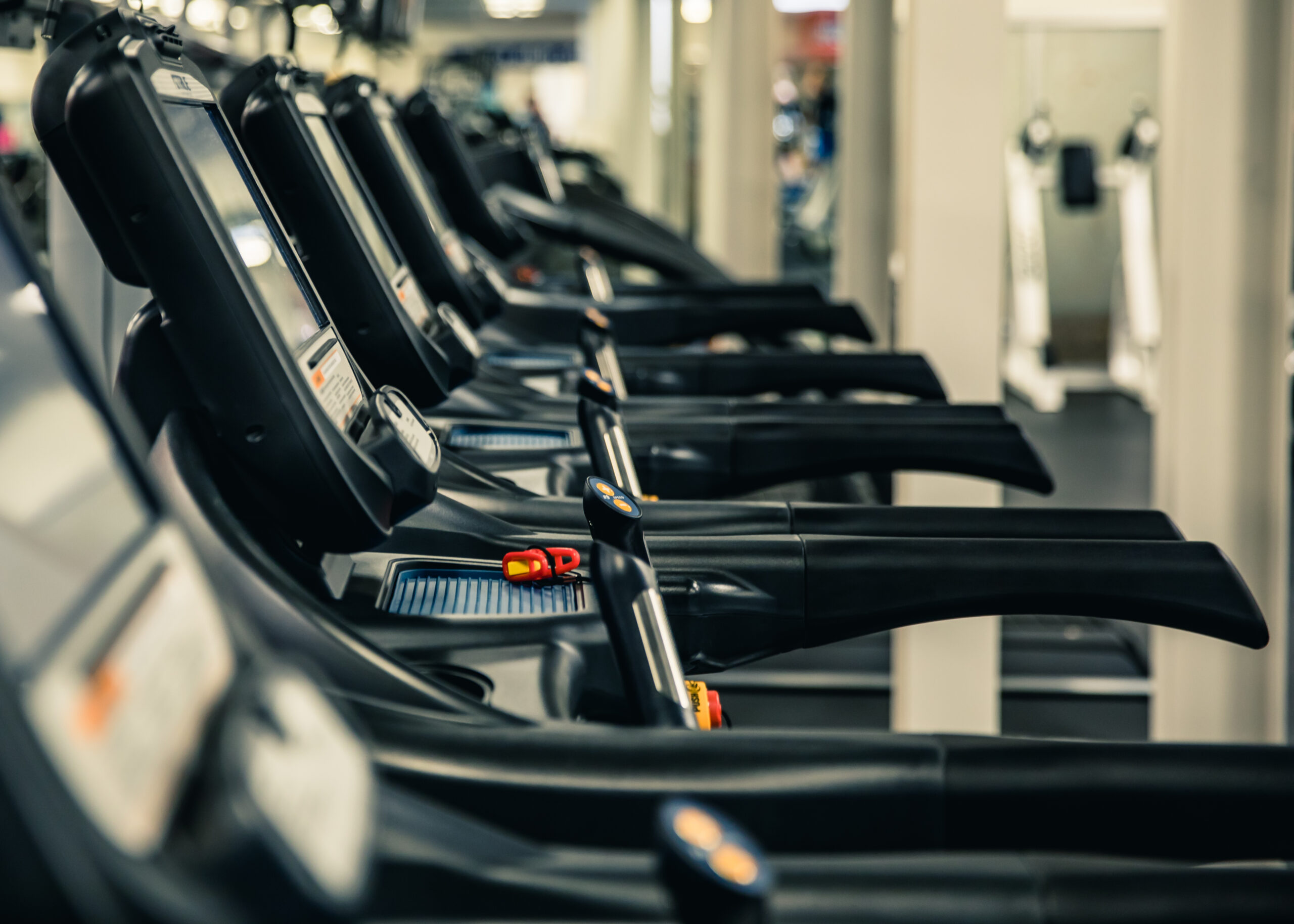 A row of black treadmills lined up in a gym, viewed from the side, with a blurred background of fitness equipment and neutral-colored walls.