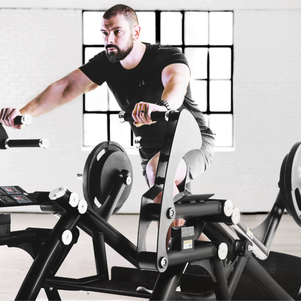 A man in a black t-shirt and shorts exercises on a stationary bike in a bright gym with large windows and modern equipment.