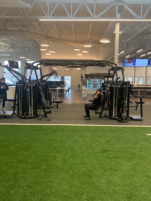 A person sits and exercises on a weight machine in a spacious gym with high ceilings, turf flooring in the foreground, and other gym equipment and people in the background.