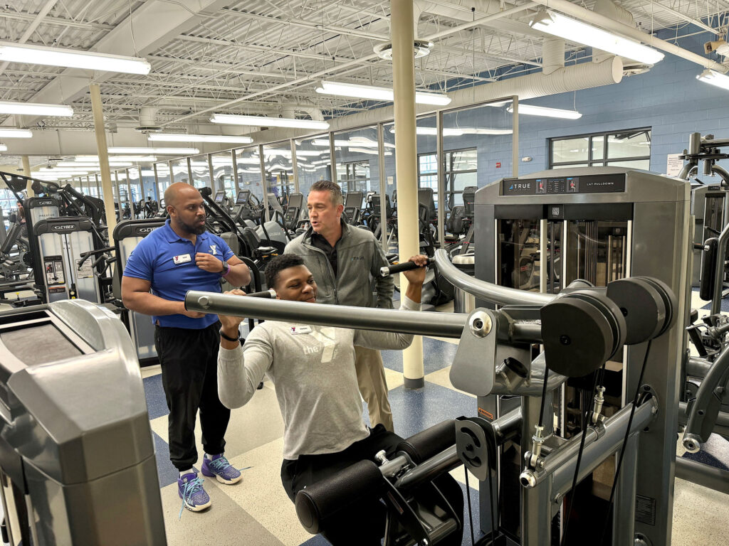 Three men are in a gym; one is seated and using a lat pulldown machine, while the other two stand nearby, observing and giving advice. The gym is spacious with various exercise equipment visible.