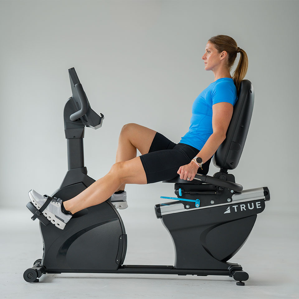 A woman in athletic clothing exercises on a recumbent stationary bike in a bright, minimalist room. She is seated upright, holding the handlebars, and pedaling with her feet strapped onto the pedals.