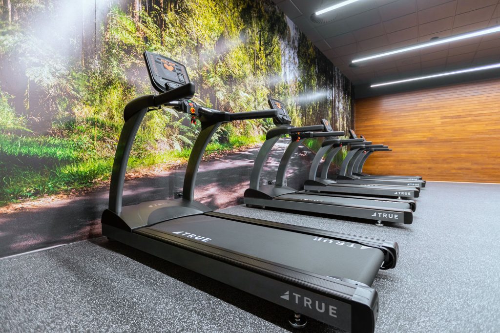 A row of four treadmills is lined up in a gym with a forest mural on one wall and wooden paneling on the other, creating a modern, nature-inspired workout space.