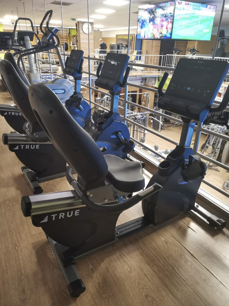 Two stationary recumbent exercise bikes with screens are positioned indoors at a gym. Several TV screens showing sports are mounted on the wall in the background, and other gym equipment is visible.