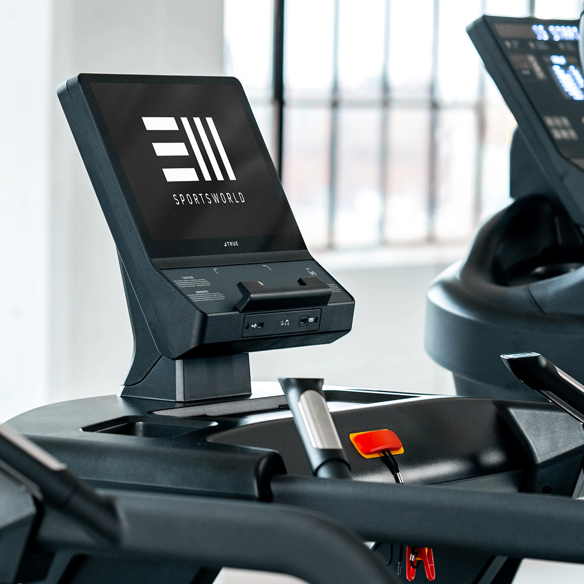 A close-up of a treadmill control panel displaying the Sportsworld logo and screen interface, with treadmill handlebars and a safety key visible in the foreground.
