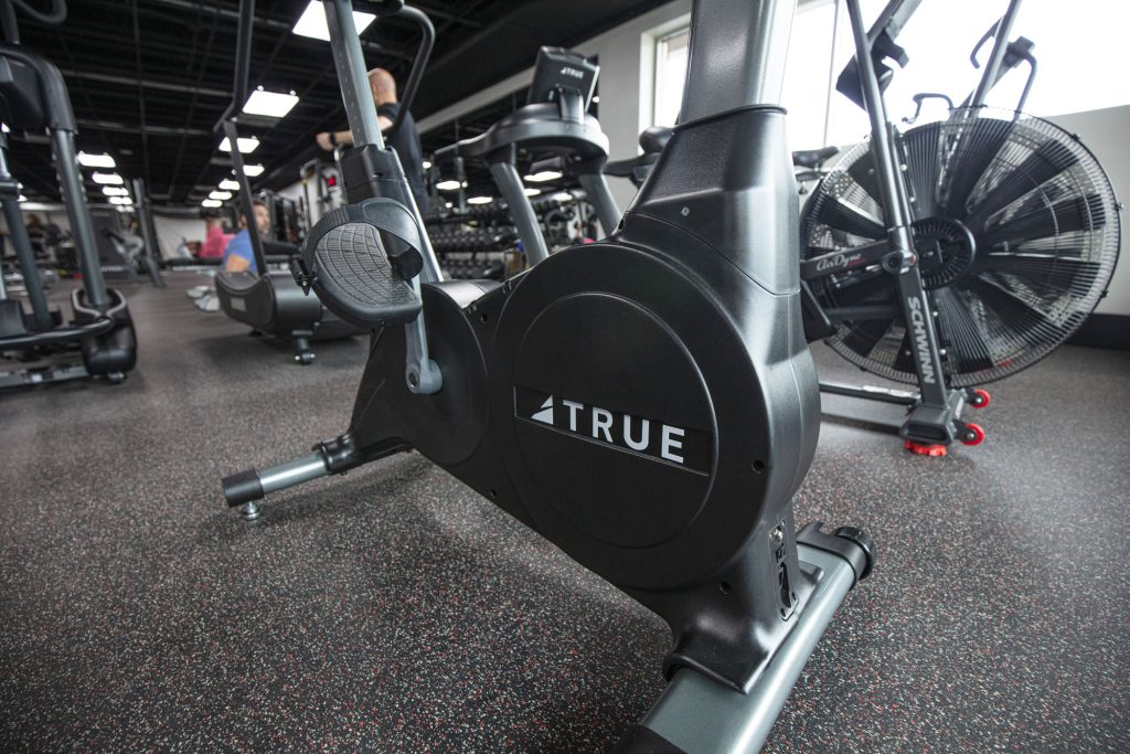 Close-up of a stationary exercise bike branded TRUE in a modern gym, with other fitness equipment and machines visible in the background.