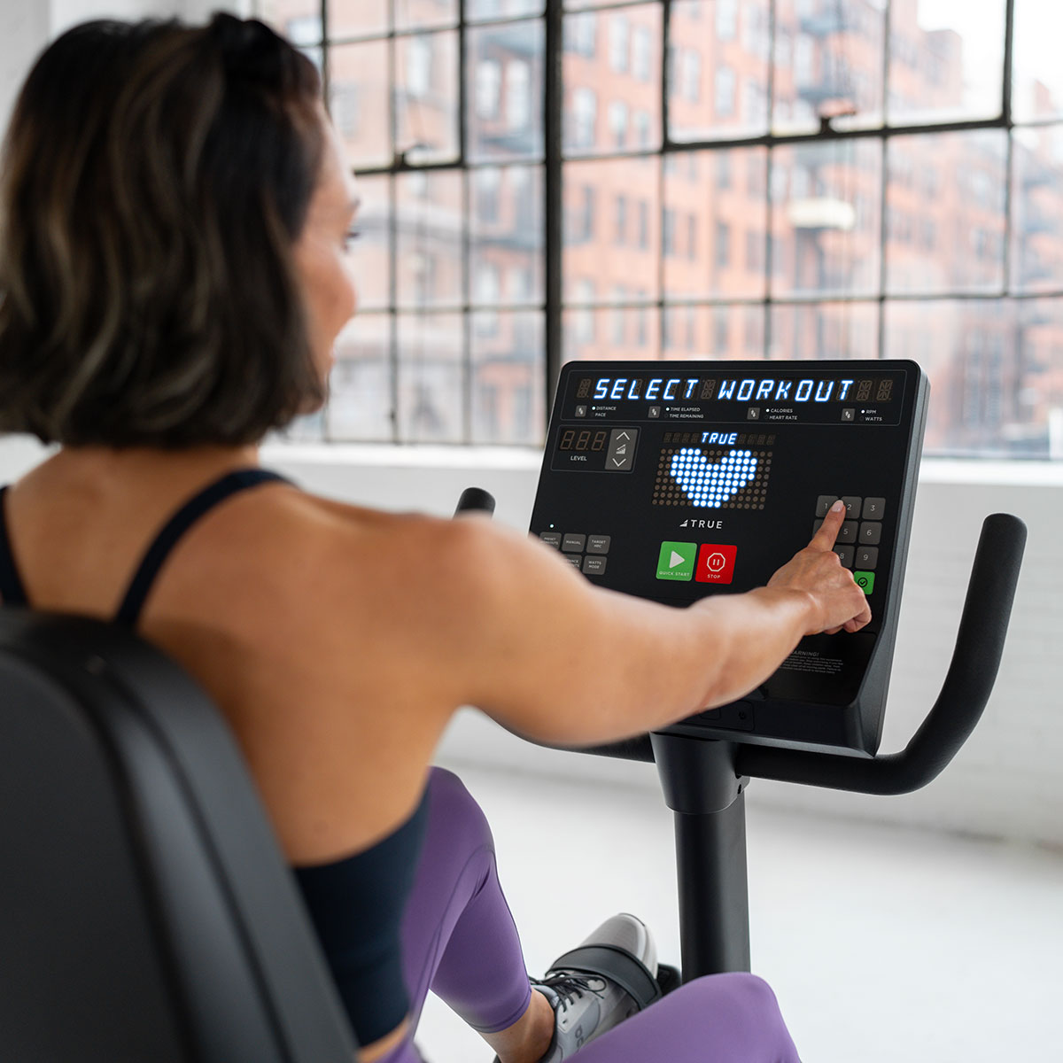 A person wearing purple leggings uses a stationary exercise bike, pressing buttons on a digital display that reads SELECT WORKOUT in a bright, modern gym with large windows overlooking city buildings.